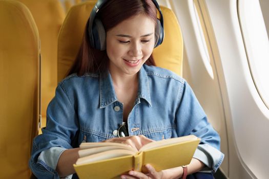 Attractive portrait of an Asian woman sitting in a window seat in economy class reading a book and listening to instrumental music during an airplane flight, travel concept, vacation, relaxation.