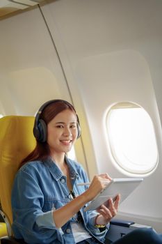 Attractive portrait of an Asian woman sitting in a window seat in economy class using tablet and listening to instrumental music during an airplane flight, travel concept, vacation, relaxation.