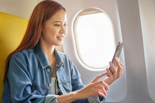 Attractive portrait of Asian woman sitting at window seat in economy class using mobile phone during inflight, travel concept, vacation, relax.