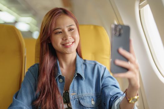 Attractive portrait of Asian woman sitting at window seat in economy class using mobile phone during inflight, travel concept, vacation, relax.