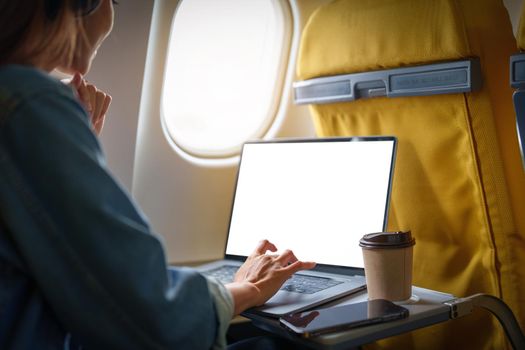 An Asian woman sits in a window seat in Economy Class using a white screen laptop computer that can use text or advertising while in flying. Travel, vacation.