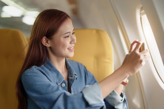 An interesting portrait of an Asian woman sitting by the window in Economy Class using a mobile phone to photograph the sky during an airplane. concept of travel, vacation, relaxation.