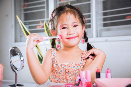 Asian adorable funny little girl making makeup her face she look in the mirror and cheek fluffy brush for powder, Learning activity to be woman, happy kid is beautiful make up with cosmetics toy