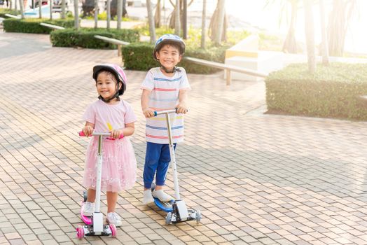 Child riding scooter. Happy Asian little kid boy and girl wear safe helmet play kick board on road in park outdoors on summer day, Active children games outside, Kids sport healthy lifestyle concept