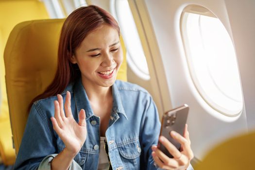 Attractive portrait of Asian woman sitting at window seat in economy class using mobile phone during inflight, travel concept, vacation, relax.