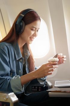 Attractive portrait of an Asian woman sitting at a window seat in economy class listening to music during a flight on a plane, travel concept, vacation, relaxation.