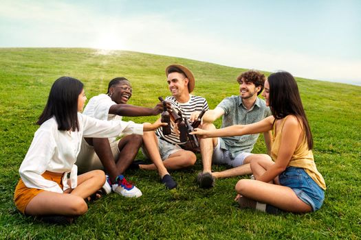 Group of multiracial happy friends having fun toasting with beers sitting on grass in public park. Friendship concept.