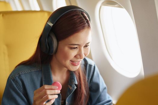 Attractive portrait of an Asian woman sitting at a window seat in economy class listening to music during a flight on a plane, travel concept, vacation, relaxation.