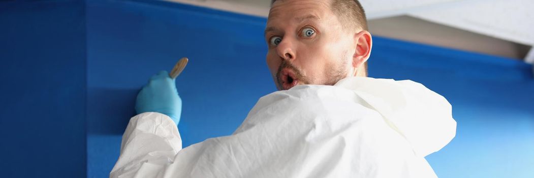 Portrait of male worker caught by surprise painting wall in blue colour. Carpenter in uniform use brush for applying paint. House design project concept