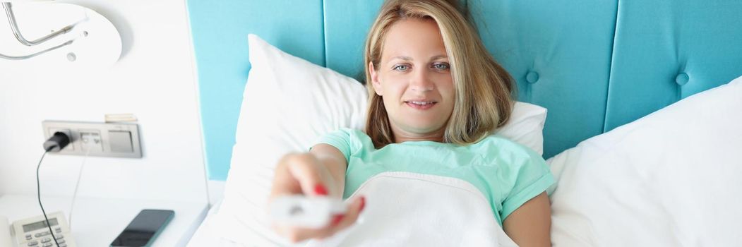 Portrait of happy woman laying in bed and use remote control to change channel on tv. Rest in hotel room, stylish interior, summer resort. Chill concept