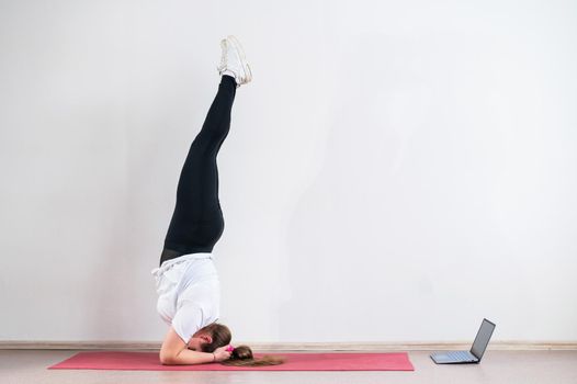 A chubby young woman watches an online yoga lesson on a laptop. Distance sports training. Forearm stand.