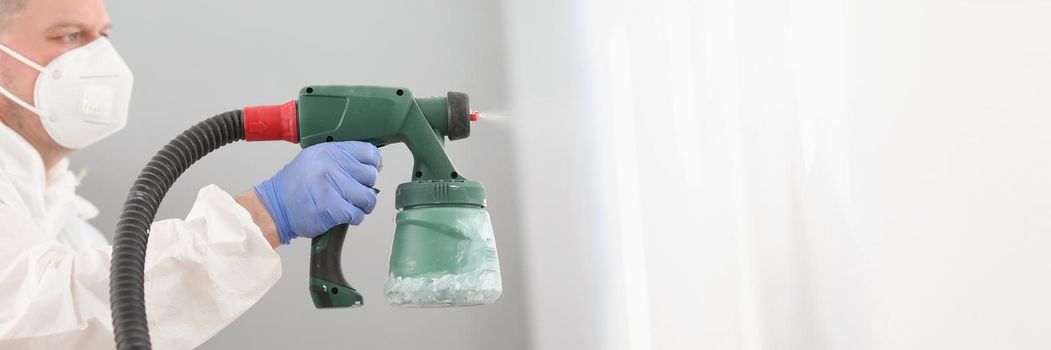 Portrait of worker using spray gun for coloring wall in white. Effective and fast way to paint walls. Renovation, interior design, household repair concept