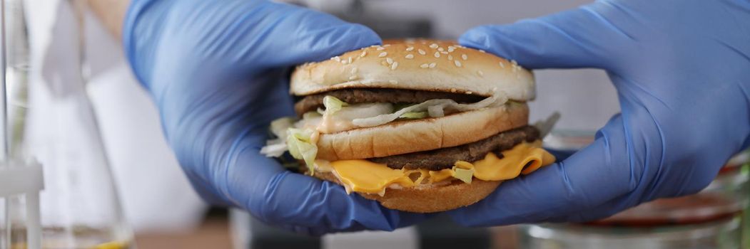 Close-up of male chemist holding tasty burger in hand with blue protective glove. Lab experiment on fastfood product. Health study and testing food concept