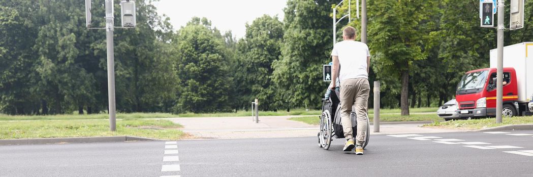 Low angle of man drive woman on wheelchair across street at pedestrian crossing. Caregiver walk with patient. Disabled people, accident, recovery concept