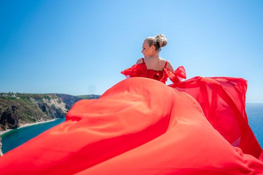 Blonde with long hair on a sunny seashore in a red flowing dress, back view, silk fabric waving in the wind. Against the backdrop of the blue sky and mountains on the seashore