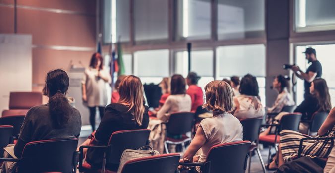 Business and entrepreneurship symposium. Female speaker giving a talk at business meeting. Audience in conference hall. Rear view of unrecognized participant in audience.