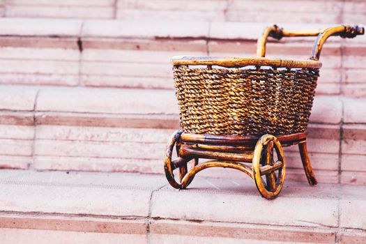 a strong open vehicle with two or four wheels, typically used for carrying loads and pulled by a horse. Wooden wicker brown cart on pink steps.