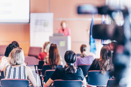 Business and entrepreneurship symposium. Female speaker giving a talk at business meeting. Audience in conference hall. Rear view of unrecognized participant in audience.