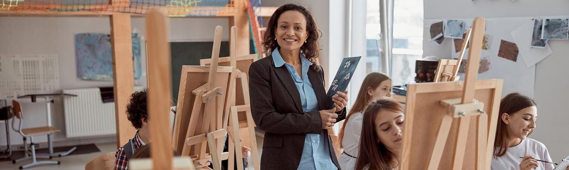 Portrait of smiling female arfican american teacher in modern classroom with kids drawing lesson in background.