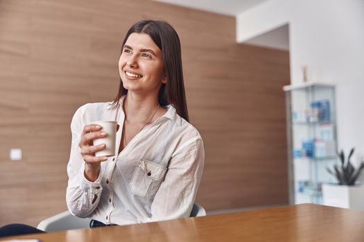 Joyful caucasian lady is drinking a coffee from a white paper cup while sitting on reception