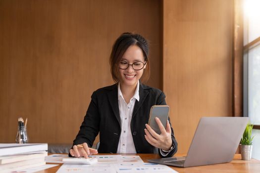 Asian woman holding smart phone while using calculator for business financial accounting calculate money bank loan rent payments manage expenses finances taxes doing paperwork concept, close up
