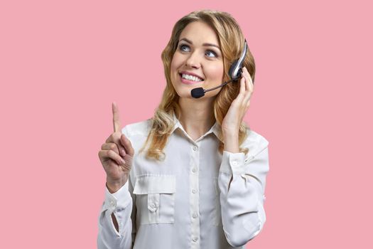 Female phone operator in headset pointing with finger upwards on pink background. Call center operator talking with a client.