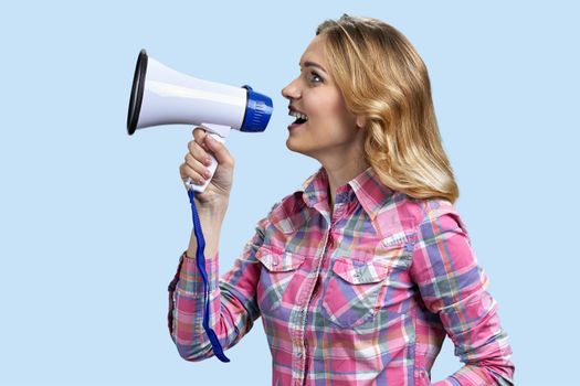 Young happy woman speaking with megaphone on light blue background. Side view of pretty girl talks into megaphone.
