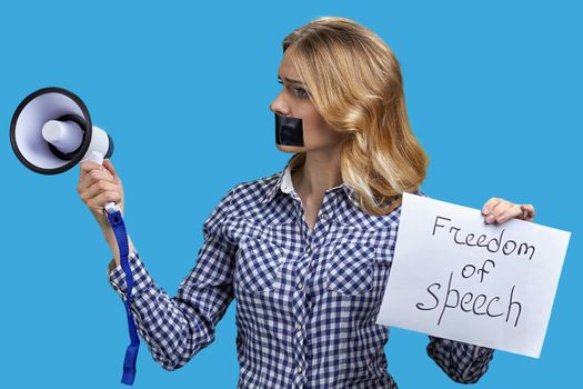 Young speechless woman with adhesive tape on mouth unable to speak in megaphone standing on color background. Freedom of speech.