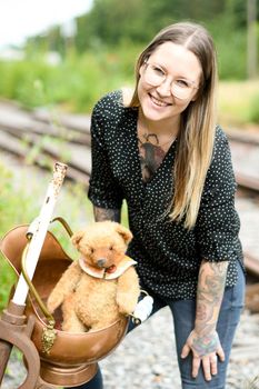 a young caucasian girl with glasses and tattoos smiles, a brown teddy bear sits in a copper pot against the background of an abandoned railway and old rails. High quality photo