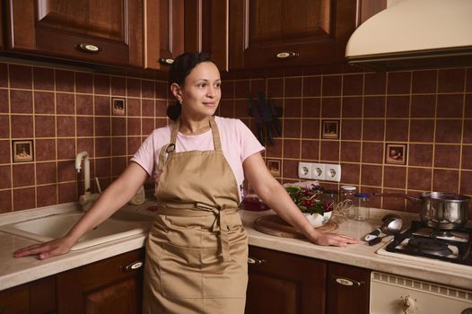 Charming middle-aged multiethnic woman housewife, confectioner wearing a chef apron, stands at kitchen countertop and smiles looking aside in the home kitchen interior