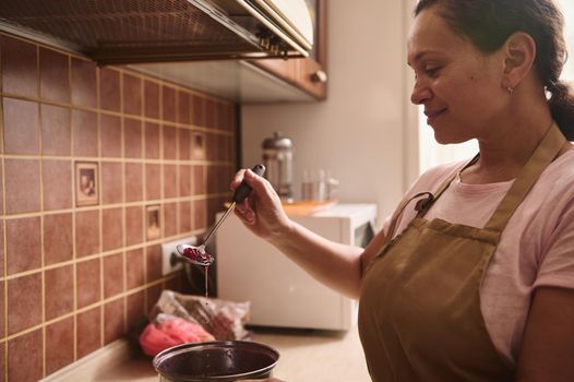 Adorable multiethnic dark haired woman wearing beige chef apron, housewife, pastry chef, standing by the stove and preparing delicious homemade cherry jam in a saucepan in a rustic wooden home kitchen
