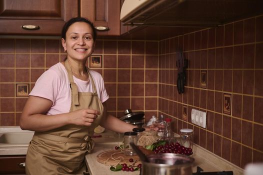 Multiethnic charming pretty woman housewife wearing a chef apron, stands at the kitchen countertop with sterilize glass jars for filling with cherry jam, preparing preserves for winter season