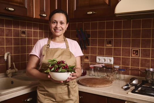 Smiling woman housewife looks at camera, standing at cozy home wooden kitchen and holding a bowl full of freshly picked cherry berries. Sterilized jars on a kitchen countertop on the background