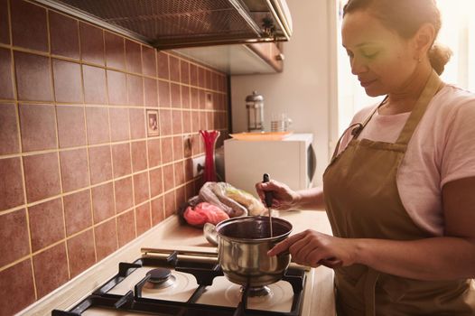 Side view of a housewife wearing a chef apron, confectioner mixing the boiling cherry jam in the saucepan, standing by stove. Sunbeams falling through large windows in the home kitchen interior