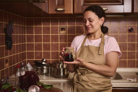 Cheerful African American woman, a pleasant housewife wearing a chef apron, holding a homemade cherry berry jam jar with the burlap and cherries on the lid, standing at cozy home kitchen interior