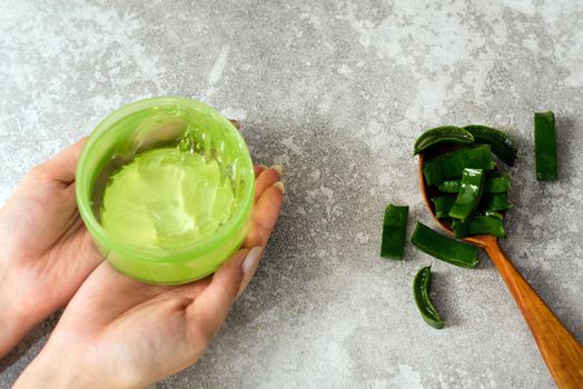 Female hands with aloe gel and a spoon with aloe leaf slices on a gray background. Making natural cosmetics at home.