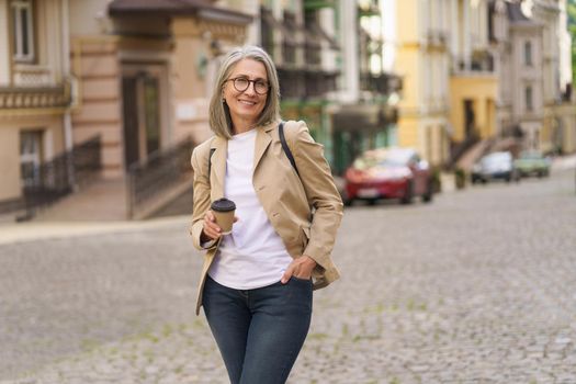 Mature businesswoman in casual formal hold disposable cup with hot coffee standing outdoors of old town. Mature woman spend free time traveling in european city enjoying cup of tea, coffee.