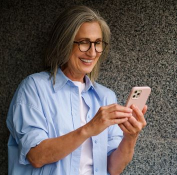 Charming mature grey hair woman talking on video call or reading text message standing outdoors leaned on marble wall happy smiling. Standing outdoors positive gorgeous woman in casual blue shirt.