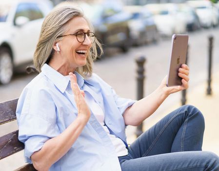 Having a video call using digital tablet and earphones mature woman with grey hair sitting on the bench at the streets of old european town. Mature woman in denim and blue shirt outdoors.