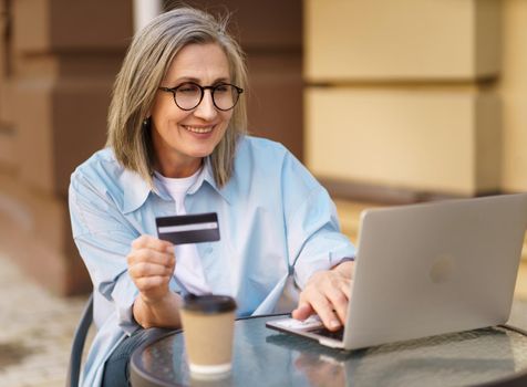 Charming mature european woman shopping or make online payment using laptop sitting outdoors street cafe terrace holding debit, credit card in hand. Mature woman happy vacation time traveling.