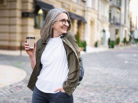 Businesswoman hold disposable cup with hot coffee. Mature woman spend free time traveling in european old town enjoying cup of tea, coffee. Grey haired woman in casual with backpack.