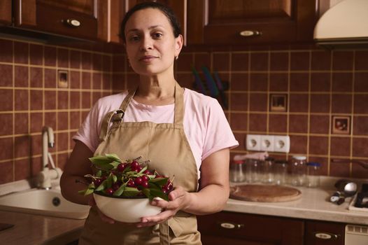 Serene charming multiethnic woman looks at camera, standing at home wooden kitchen and holding a bowl full of freshly picked cherry berries. Sterilized jars on a kitchen countertop on the background