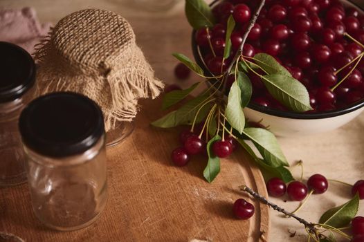 Close-up. Top view of sterilized glass jam jars with lids on a wooden board next to an enameled bowl of freshly picked cherries on a kitchen countertop. Preparation for conservation