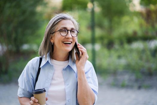 European gray haired businesswoman talking on the phone while drinks coffee on the go using paper cup in city garden or park. Mature woman at coffee break, wearing blue shirt and white t-shirt.