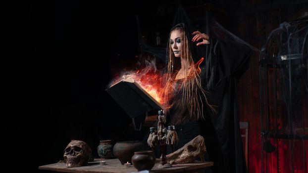 Halloween concept. Witch dressed black hood standing dark dungeon room use magic book conjuring magic fire spell. Female necromancer wizard gothic interior with skull, cage, spider web