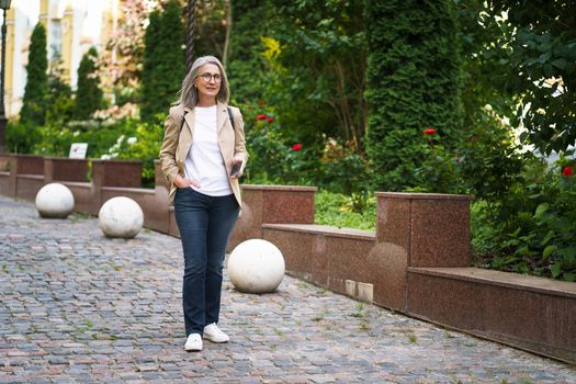 Beautiful mature business woman in casual formal with phone in hand walking on street of old town from work or back to office. Mature woman spend free time traveling in european city.