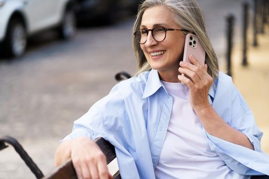 Enjoying summer time mature woman with grey hair talking on the phone holding smartphone sitting on the bench at the streets of old european town. Mature woman answering call outdoors.