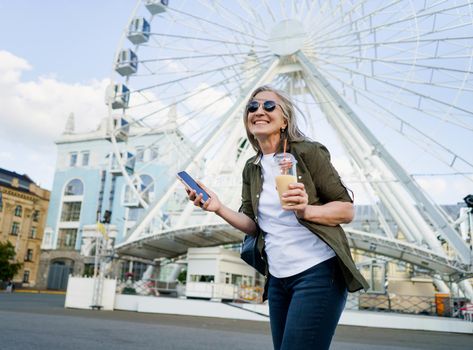 European charming grey haired woman happy enjoying free time after work or traveling, holding phone while having juice on the go using plastic cup in city and ferris wheel on background.