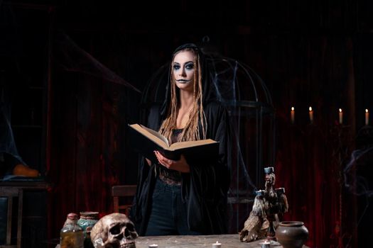 Halloween concept. Witch dressed black hood with dreadlocks standing dark room use magic book conjuring magic spell. Female necromancer wizard gothic interior with skull, cage, spider web Occultism