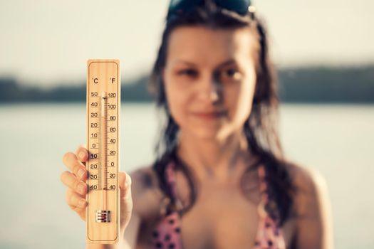 Beautiful young woman with a thermometer in her hand on the background of the lake. Summer vacation and heat concept, selective focus.